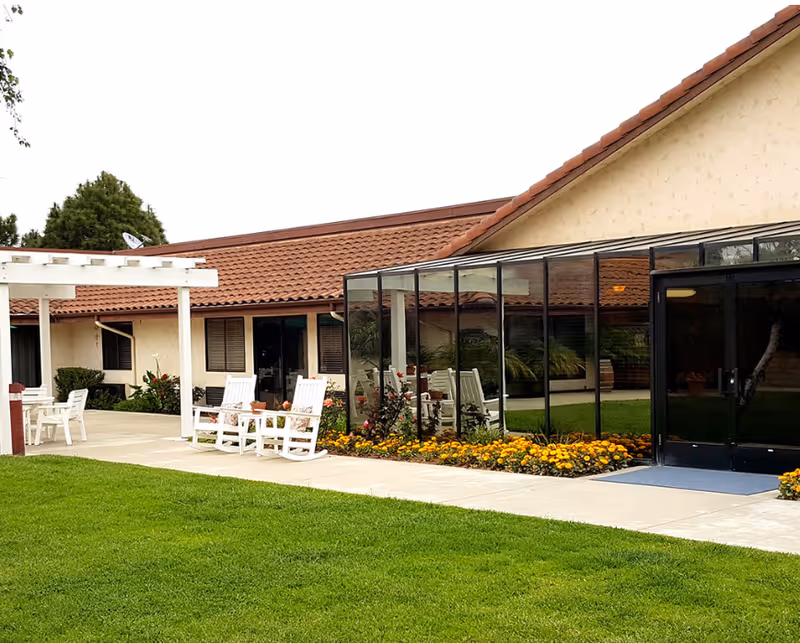 Outdoor view of a senior living facility with a well-maintained green lawn, a concrete walkway, white rocking chairs, a white pergola, and a building with beige walls and a red tiled roof. There is a glass-enclosed patio area with flowers planted along the edge.