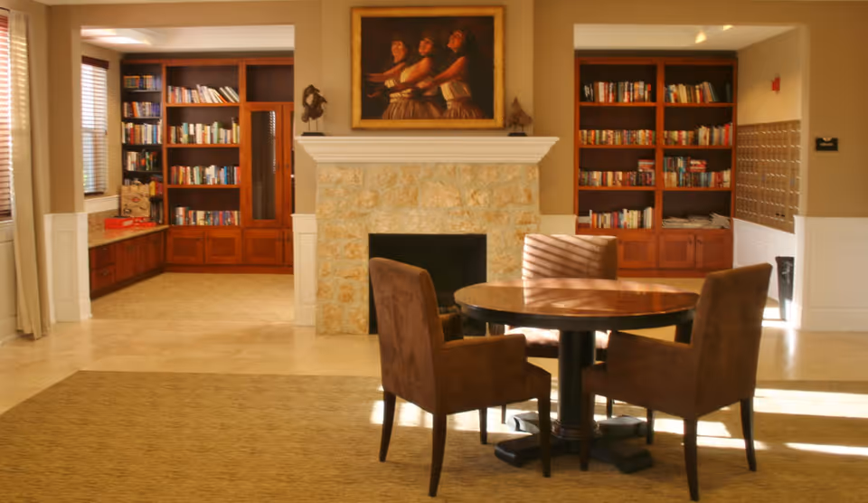 A cozy interior common area featuring a round wooden table with four brown upholstered chairs. Behind the table is a stone fireplace with a painting of three women above it. On either side of the fireplace are built-in wooden bookshelves filled with books. The room is warmly lit with natural light coming through windows with blinds.