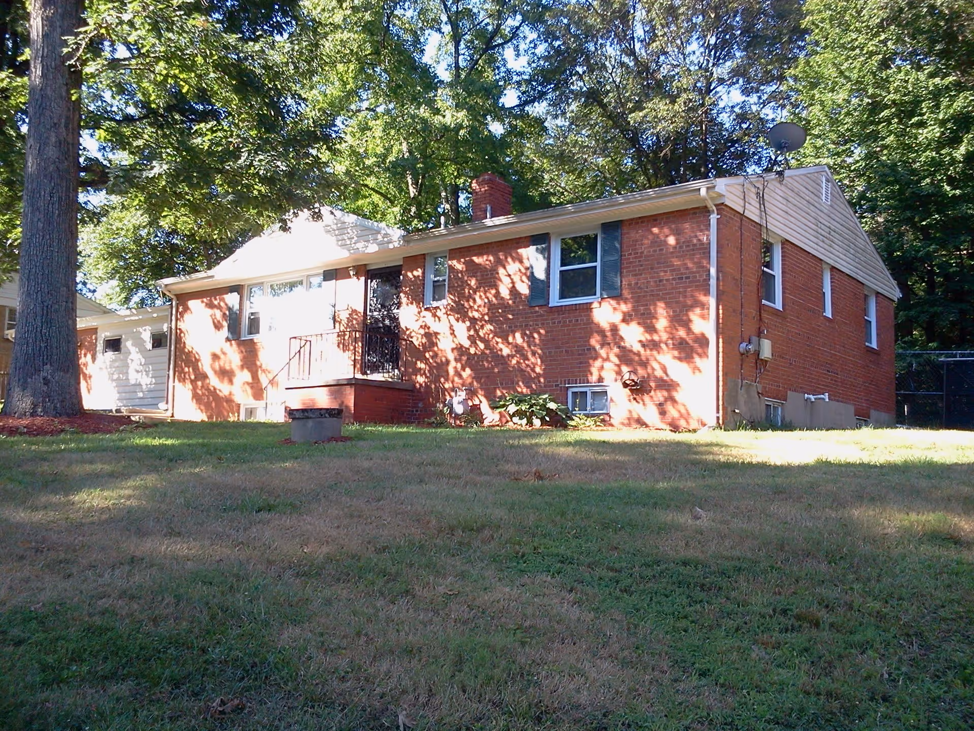 A single-story brick house with a small front porch and black metal railing, surrounded by green grass and large trees casting shadows on the building.