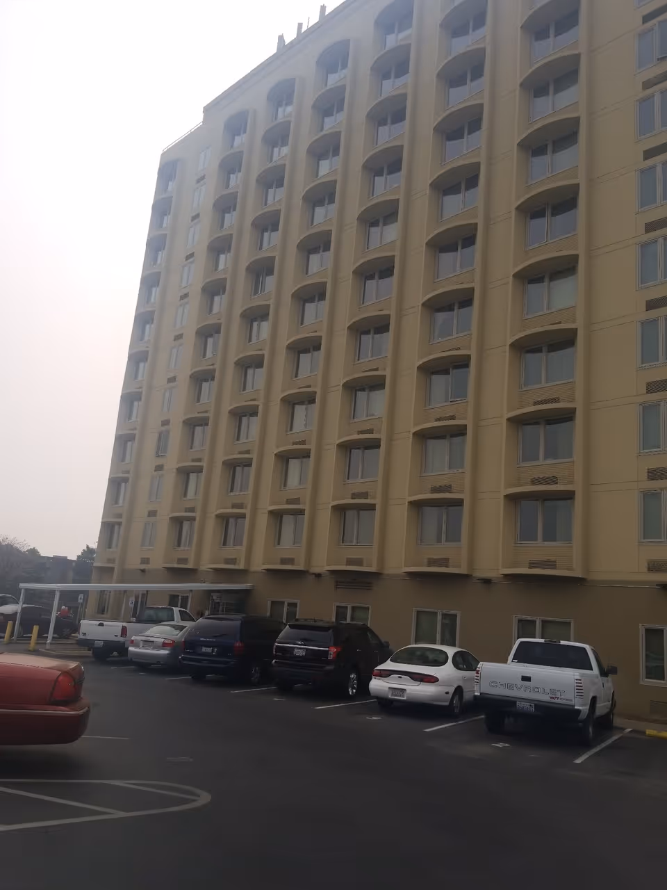 Tall multi-story beige building with repeating arched windows and a parking lot with several cars in front.