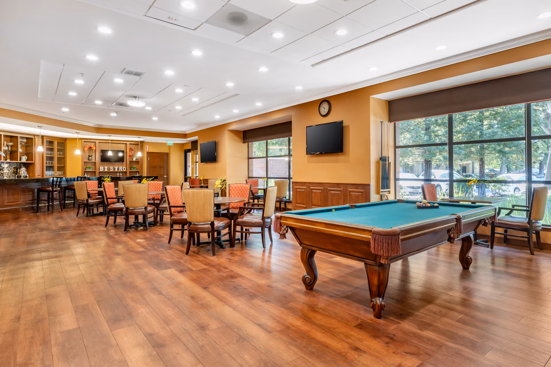 A spacious senior living common area with a pool table in the foreground, multiple tables and chairs arranged for dining or socializing, large windows letting in natural light, two wall-mounted televisions, and a bar area labeled 'Bistro' in the background.
