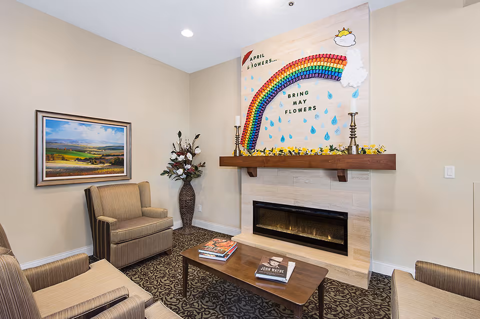 A cozy living room area with a modern fireplace set in a tiled wall. Above the fireplace is a colorful rainbow decoration with the words 'April Showers... Bring May Flowers' and a small sun and cloud illustration. The room has beige walls, a patterned carpet, a wooden coffee table with books on it, two striped armchairs, a framed landscape painting on the wall, and a tall vase with flowers in the corner.