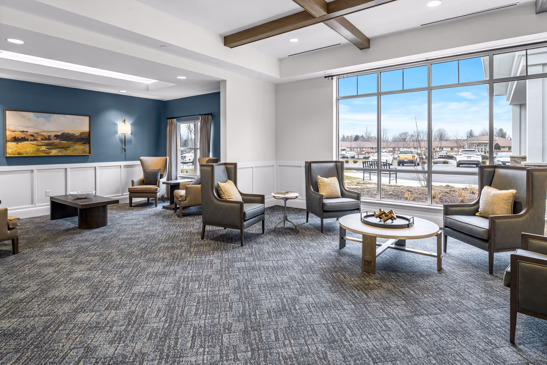 A spacious and well-lit living room area with multiple armchairs arranged around two coffee tables. The room features large windows allowing natural light to fill the space, a blue accent wall with a landscape painting, and a carpeted floor. Outside the window, parked cars and a bench are visible.