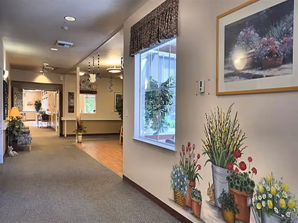 Interior hallway of an assisted living facility with a painted potted-plant mural on the right wall, a window with a 'welcome' decal, and seating and lamps down the corridor.