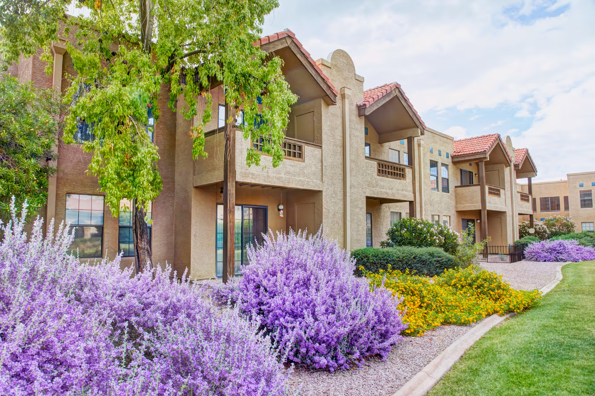 Exterior view of a senior living facility building with beige stucco walls and red tile roofs. The building has balconies and large windows. In the foreground, there are well-maintained landscaping features including vibrant purple and yellow flowering bushes, green shrubs, and a tree with green leaves. The sky is partly cloudy.