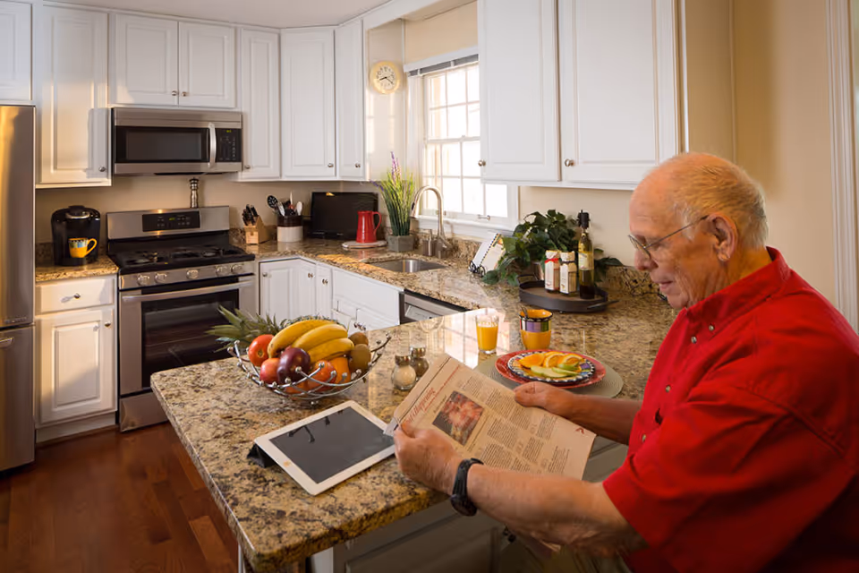 An elderly man in a red shirt is sitting at a kitchen counter reading a newspaper. The kitchen has white cabinets, granite countertops, a stainless steel stove, microwave, and refrigerator. On the counter, there is a bowl of assorted fruits, a tablet, a plate with sliced fruit, a glass of orange juice, and various kitchen items near the window.