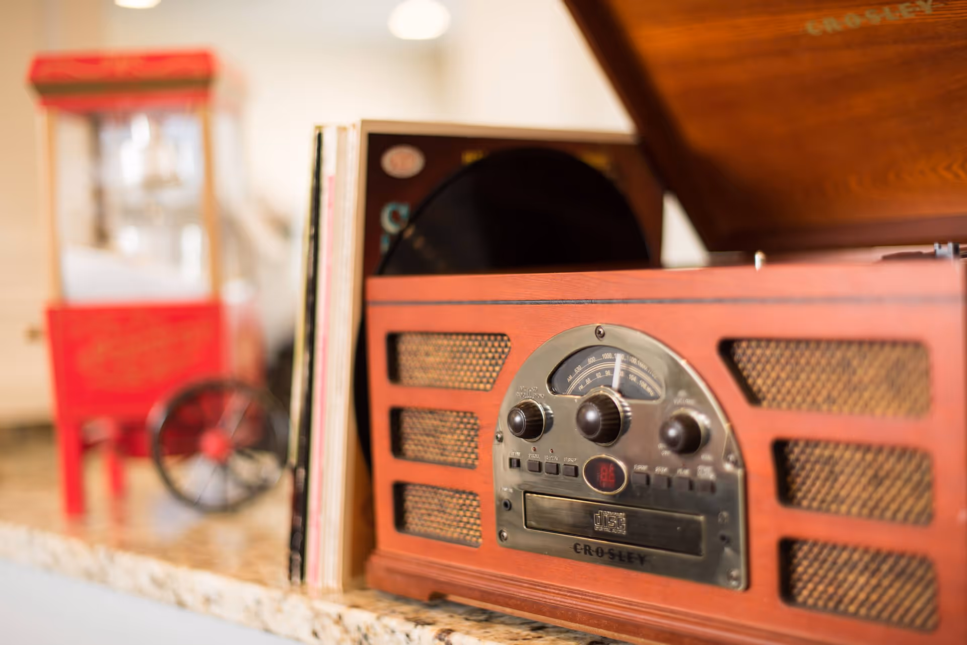 Close-up of a vintage Crosley record player and vinyl records on a countertop with a small red popcorn machine blurred in the background.