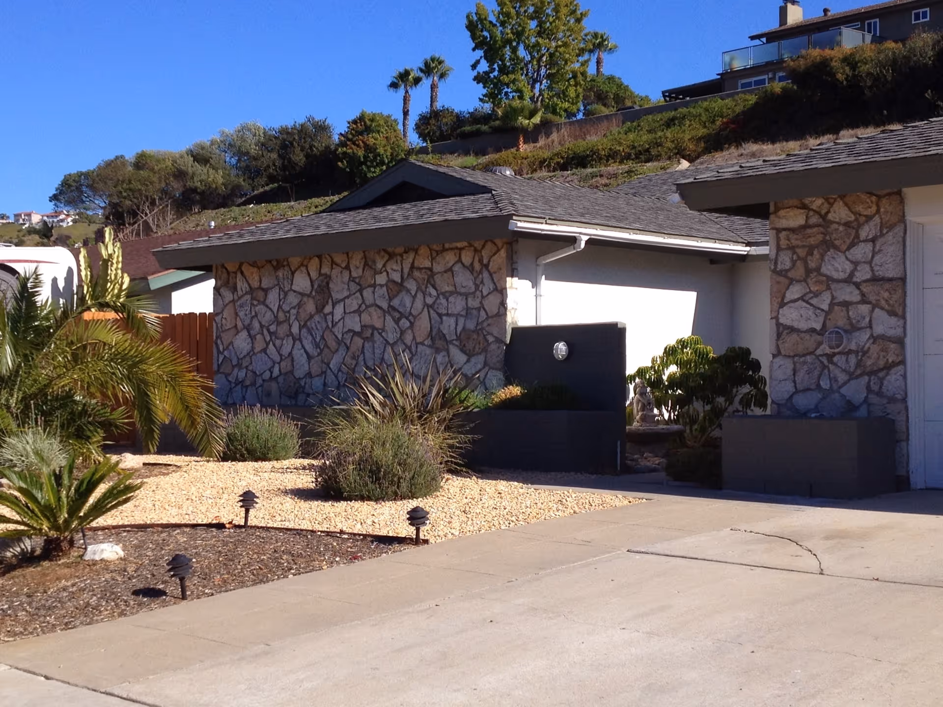 Exterior view of a single-story building with stone facade walls, a driveway, and landscaped front yard featuring small palm trees, bushes, and gravel. The sky is clear and blue with some trees and buildings visible in the background.