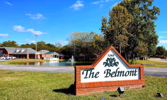 Outdoor view of The Belmont Assisted Living facility showing a brick sign with the name 'The Belmont' and the number 7295. In the background, there is a pond with a water fountain, trees, a clear blue sky, and a building with a parking area and cars.