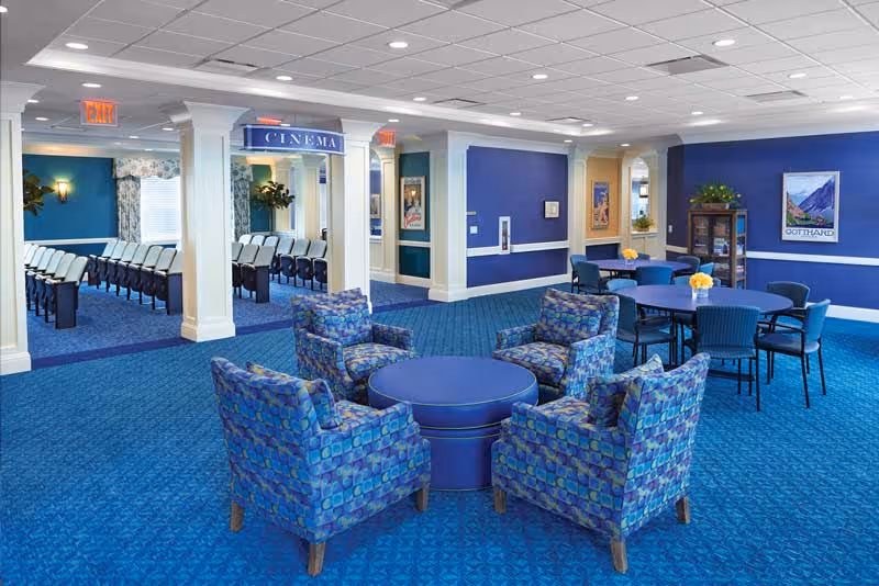 Communal lounge in an assisted living facility with blue patterned armchairs around a round table, additional dining tables, and a small cinema entrance.