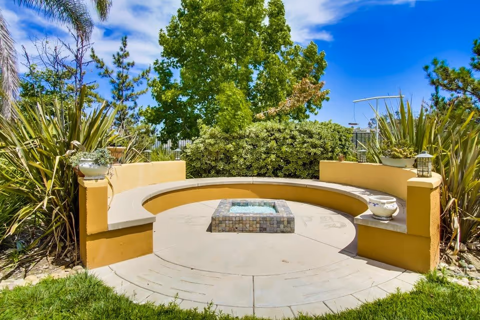 Curved yellow stucco bench surrounding a small tiled water feature in a sunny garden courtyard with trees and plants.