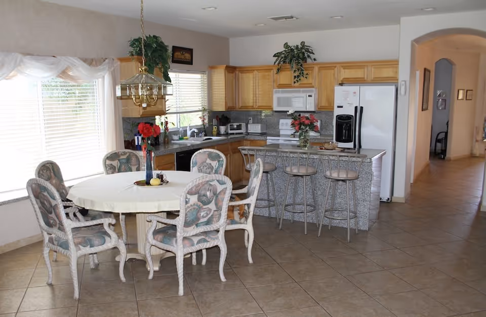 A bright kitchen and dining area with a round table covered with a white tablecloth and six cushioned chairs with patterned upholstery. The kitchen features wooden cabinets, a white refrigerator, a microwave, a stove, and a granite countertop island with three bar stools. There are plants and flowers decorating the space, and large windows with white blinds letting in natural light.
