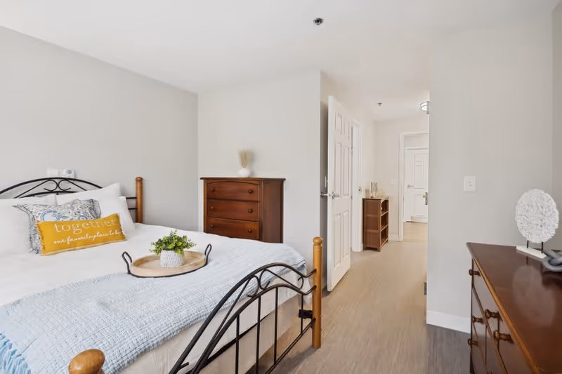A bright and clean bedroom with a bed featuring a black metal frame and wooden posts. The bed is made with white linens, a light blue throw blanket, and decorative pillows including one yellow pillow with the word 'together' on it. There is a wooden dresser with a small vase on top against the wall, and another wooden dresser with a decorative white sculpture on it. The room has light-colored walls and flooring, with an open door leading to a hallway.