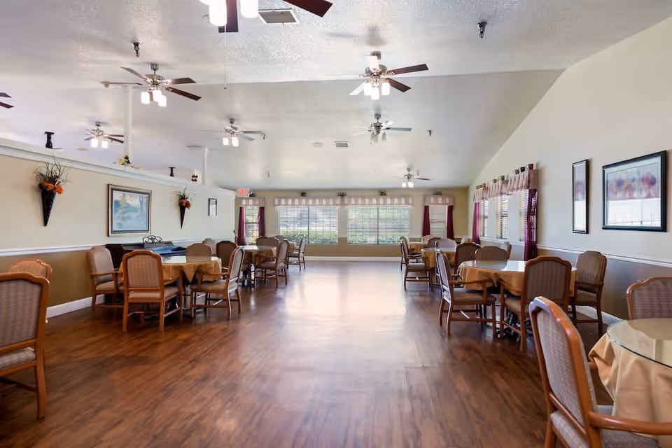 A spacious dining room with multiple round tables covered with beige tablecloths, surrounded by wooden chairs with cushioned seats. The room has wooden flooring, several ceiling fans with lights, large windows with blinds and curtains, and framed artwork on the walls. There are decorative wall-mounted flower arrangements and an exit door at the far end.