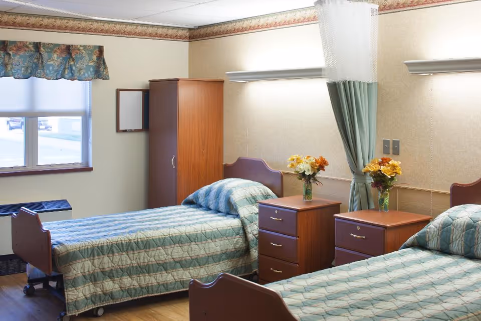 A well-lit room with two single beds, each with a green and blue patterned bedspread and matching pillows. Between the beds are two wooden nightstands with drawers, each holding a vase of yellow and orange flowers. A green privacy curtain hangs between the beds. There is a wooden wardrobe against the wall and a window with a floral valance letting in natural light.