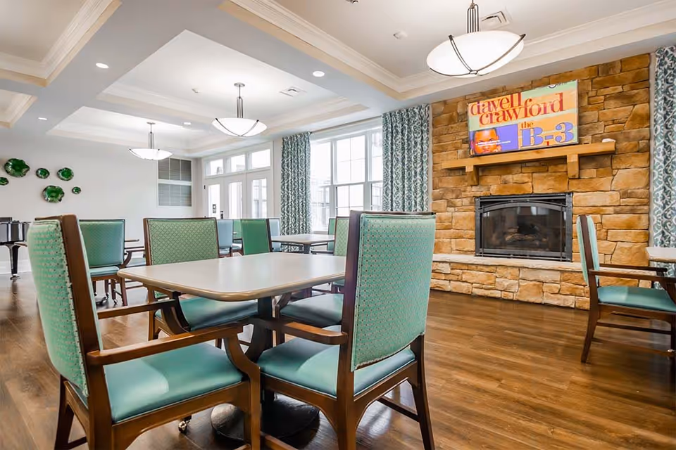 A bright and spacious dining room with multiple tables and green cushioned chairs. The room features large windows with patterned curtains, a stone fireplace with a colorful artwork above it, and modern ceiling lights. The floor is wooden, and there is a piano visible in the background.