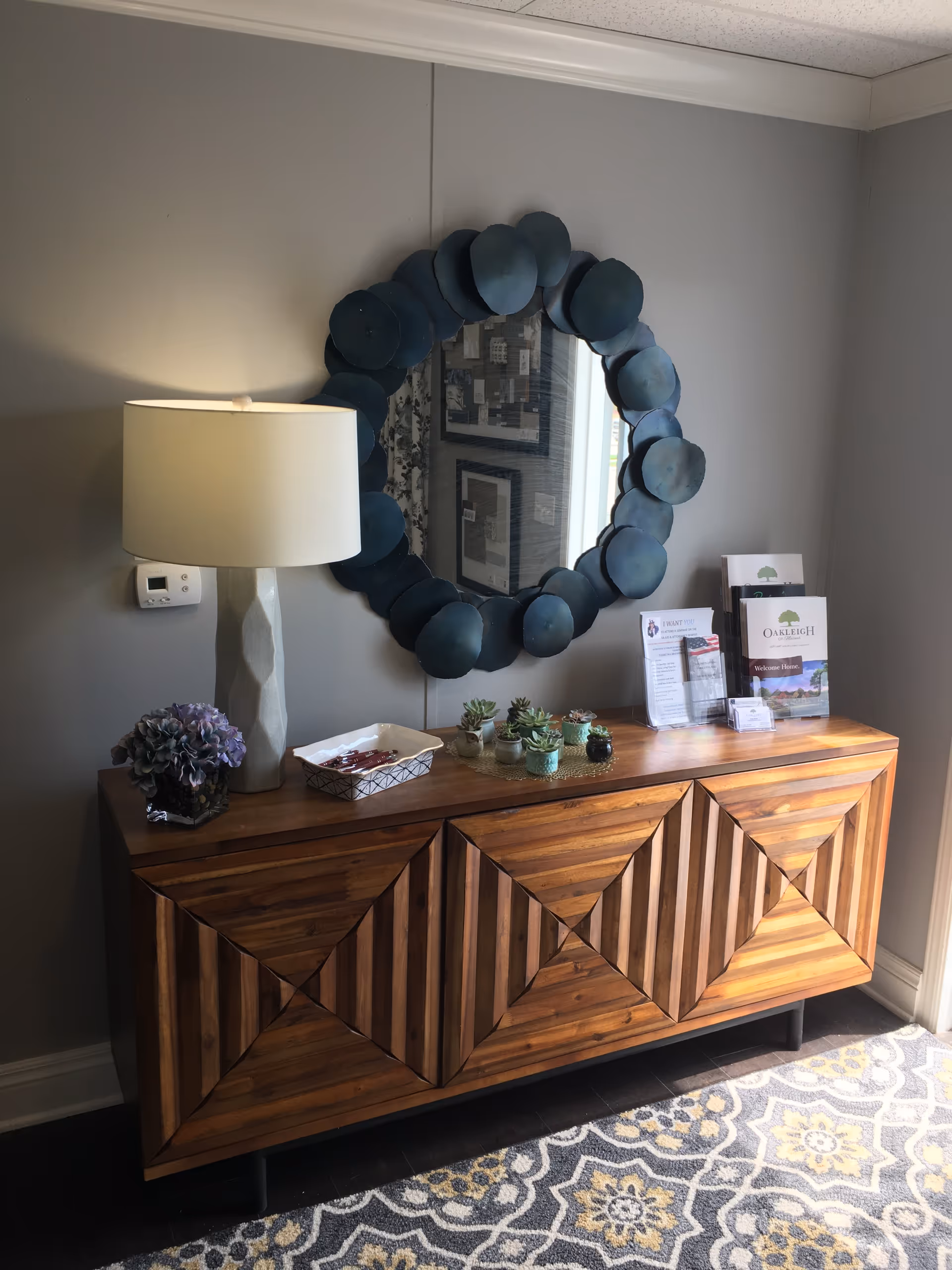 A wooden sideboard with a geometric pattern on the front, topped with a white table lamp, a small purple flower arrangement, a tray with utensils, several small potted succulents, and brochures. Above the sideboard is a round mirror with a decorative blue frame. The wall behind is painted gray and a patterned carpet is visible on the floor.