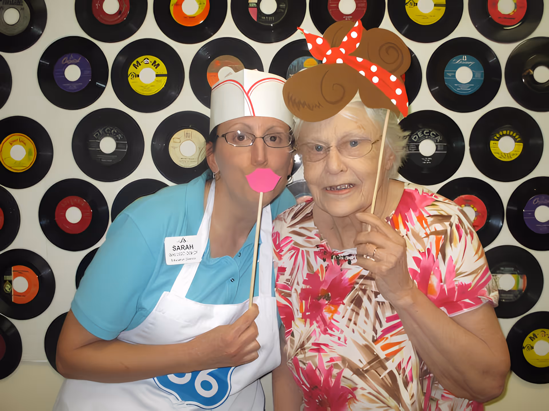 Two women posing in front of a wall decorated with vinyl records. One woman is wearing a blue shirt, white apron, and a paper hat, holding a stick with pink lips in front of her face. The other woman is wearing glasses and a floral shirt, holding a stick with a cartoonish hair and red polka dot headband prop in front of her head.