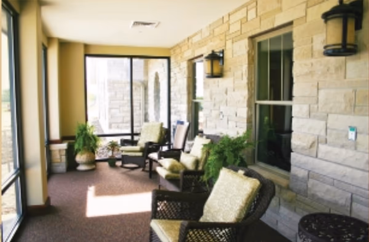 A sunlit indoor seating area with cushioned wicker chairs and potted plants along a stone wall with windows and wall-mounted lanterns.