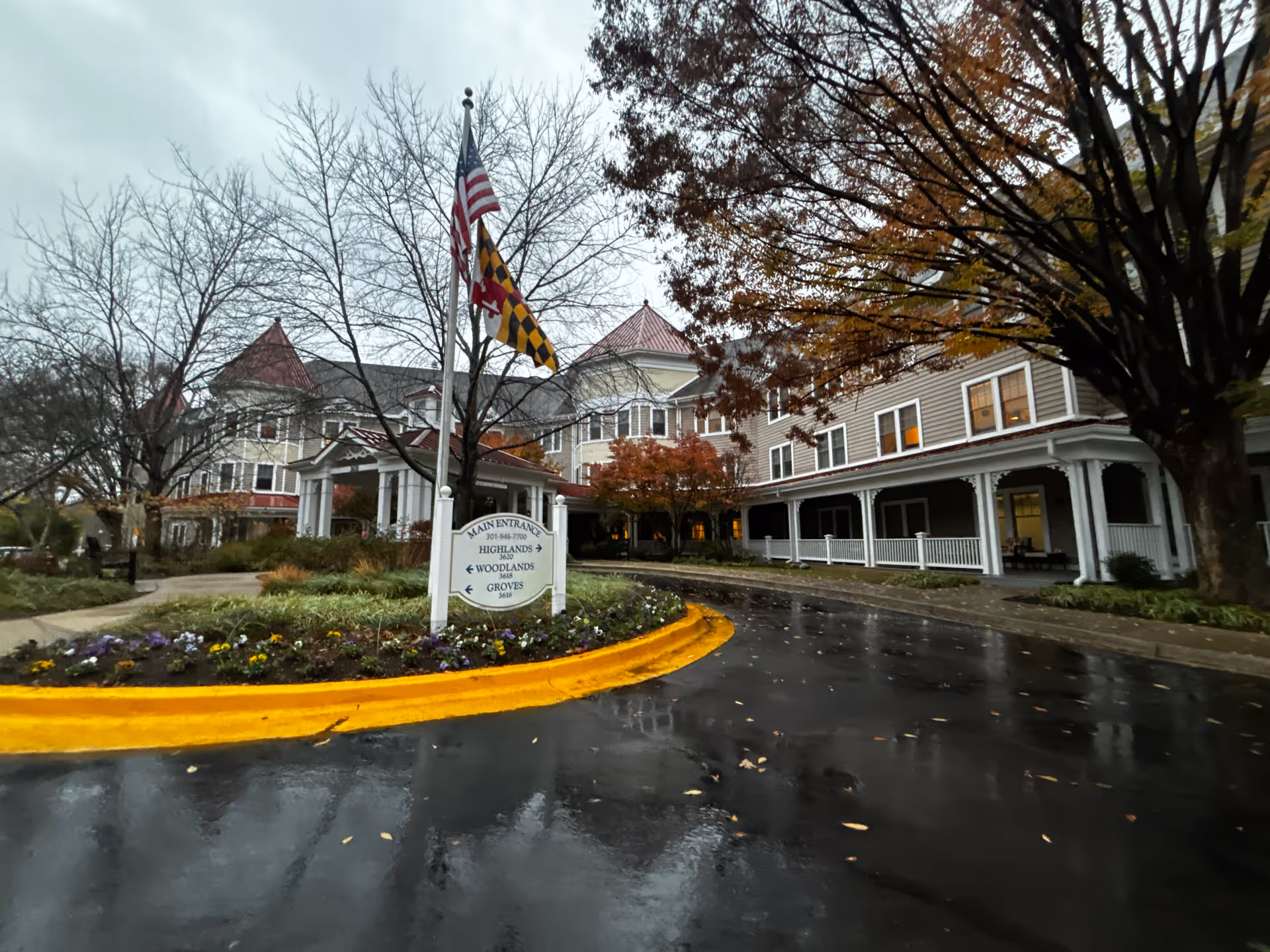Front entrance of a senior living building with flags, landscaping, and a wet circular driveway under an overcast sky.