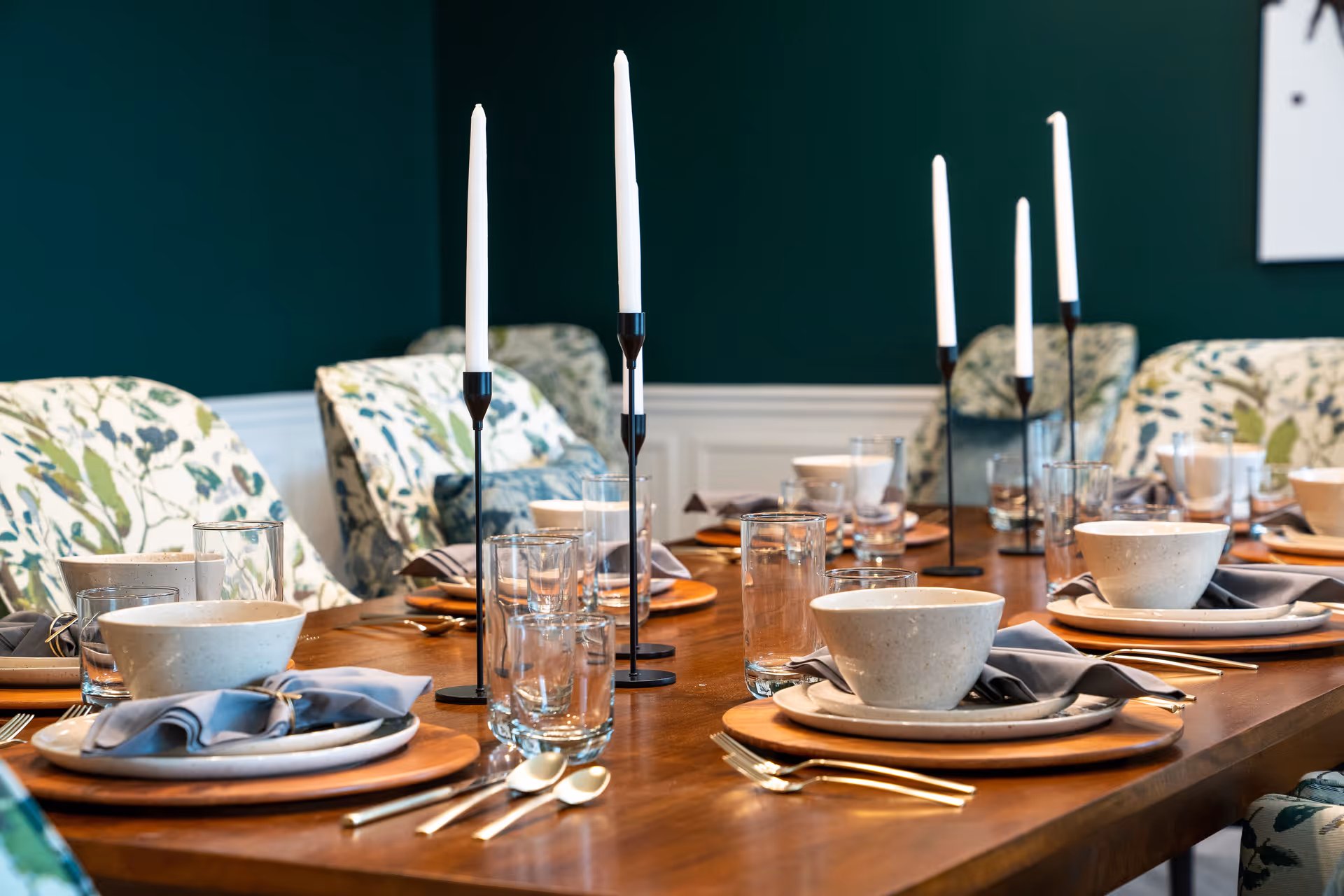 A wooden dining table set with ceramic bowls, plates, glasses, and silverware. The table is decorated with tall white candles in black candle holders. The chairs around the table have floral patterned upholstery, and the background wall is painted dark green with white wainscoting.
