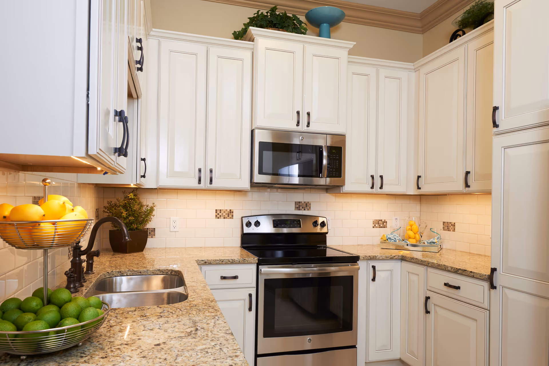 A modern kitchen with cream-colored cabinets, granite countertops, a stainless steel oven and microwave, a double sink with a dark faucet, and bowls of lemons and limes on the counter. There are decorative plants and a blue vase on top of the cabinets.