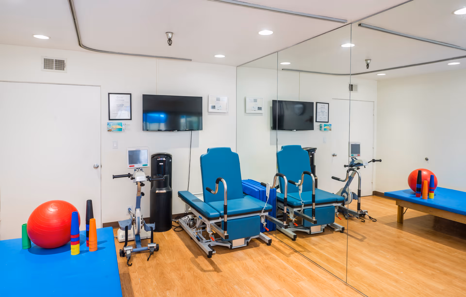 A physical therapy room with two blue therapy chairs, a stationary exercise bike, a large mirror covering one wall, a blue padded table with a red exercise ball and colorful cones, and a wall-mounted TV.