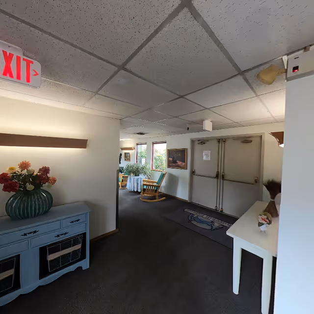 Interior hallway of a retirement community with a dark carpeted floor, white walls, and a drop ceiling. There is a blue console table with a green vase holding colorful flowers on the left, and a white table with decorative items on the right. At the end of the hallway, there are two large double doors with push bars, a rug in front of them, and a seating area with wooden rocking chairs and a small table near windows letting in natural light. An illuminated red EXIT sign is visible on the ceiling.