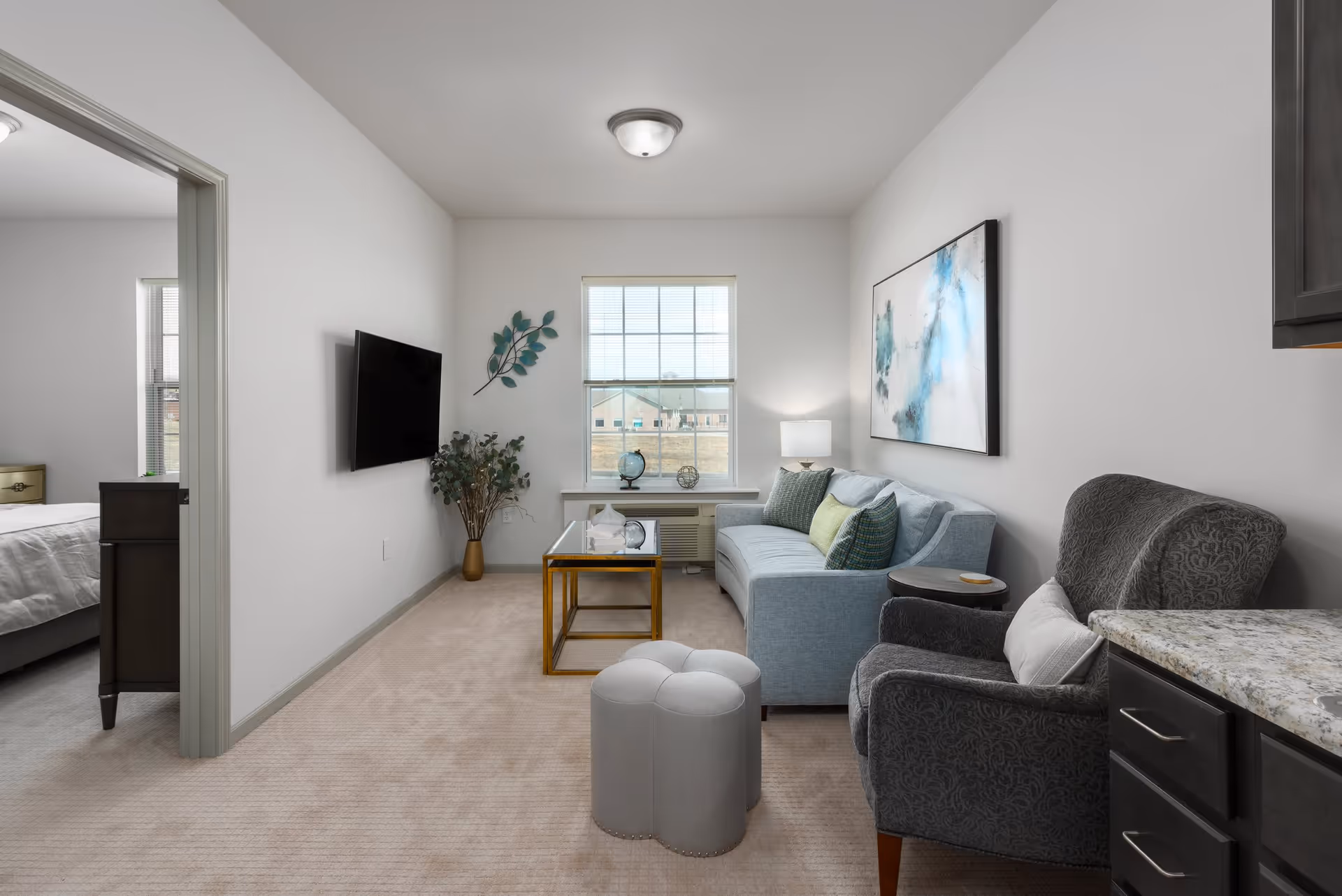 A cozy living room with a light blue sofa adorned with green and blue pillows, a gray patterned armchair with a white pillow, a small round side table, and a glass coffee table with a gold frame. A flat-screen TV is mounted on the wall opposite the sofa. There is a window with blinds letting in natural light, and a decorative plant and wall art add to the room's decor. The living room opens into a bedroom with a bed visible through the doorway.