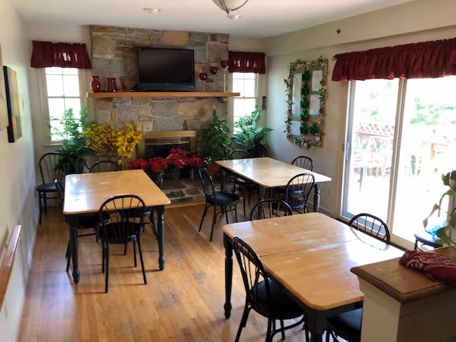 A cozy dining area with four wooden tables and black metal chairs arranged around them. The room features a stone fireplace with a mounted flat-screen TV above it, flanked by two windows with red valances. There are various plants and floral decorations around the fireplace and near a sliding glass door that leads outside. The floor is wooden, and the walls are light-colored.