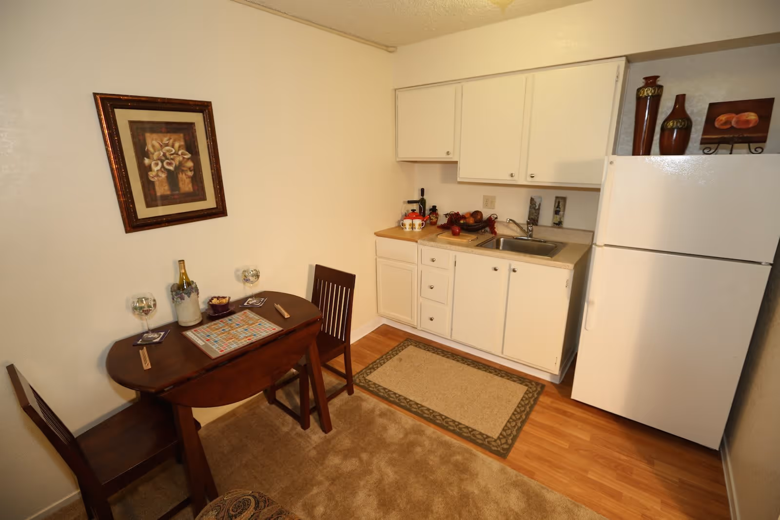A small kitchen area with white cabinets, a countertop with a sink, and a white refrigerator. Next to the kitchen is a small wooden dining table with two chairs. On the table, there is a Scrabble board game, two wine glasses, a bottle of wine in an ice bucket, and some snacks. A framed floral painting hangs on the wall above the table. The floor is a combination of wood and carpet with a small rug in front of the kitchen sink.