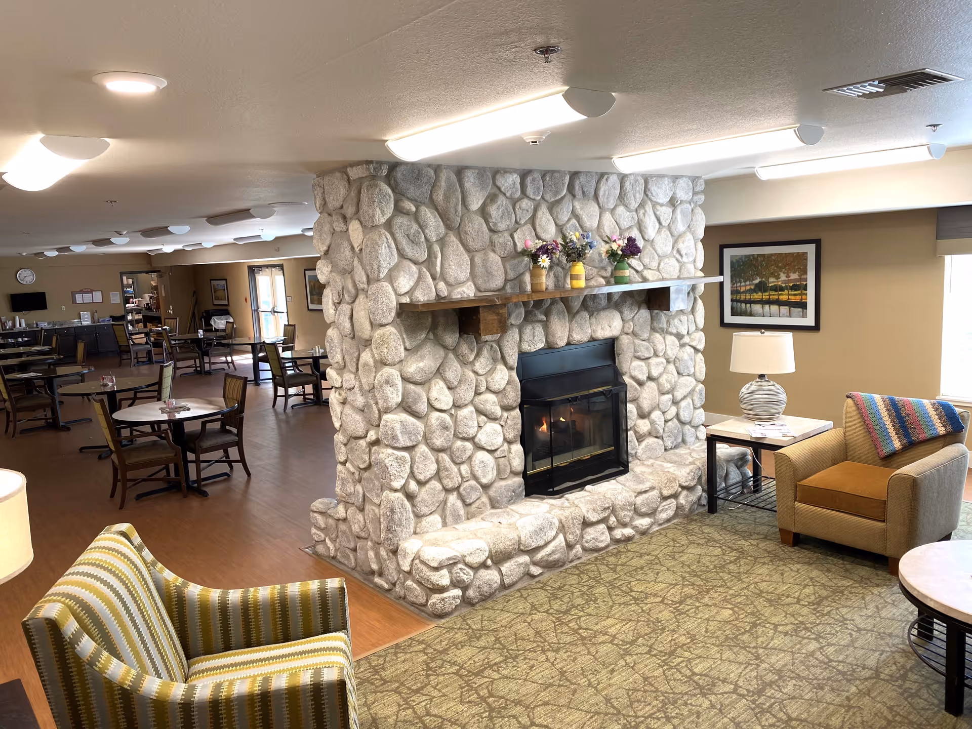 Interior view of a senior living facility common area featuring a large stone fireplace with a wooden mantel decorated with flower vases. The room has a cozy seating area with an armchair and a striped chair, a side table with a lamp, and a framed painting on the wall. In the background, there are multiple dining tables and chairs arranged on a wooden floor under ceiling lights.