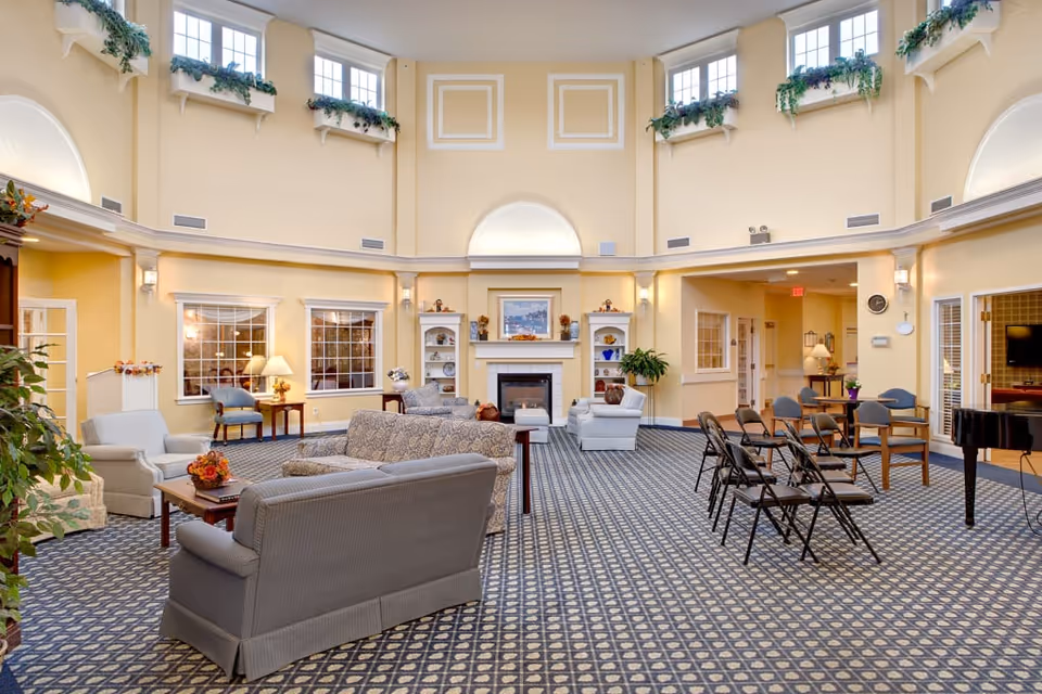 A spacious and well-lit living room area with high ceilings and multiple windows near the ceiling. The room features patterned carpet flooring, several upholstered sofas and armchairs arranged around a coffee table, a fireplace with built-in shelves on either side, and a grand piano. There are also several folding chairs and a few chairs with armrests arranged in rows. The walls are painted a soft yellow, and there are decorative plants on ledges near the ceiling and around the room.