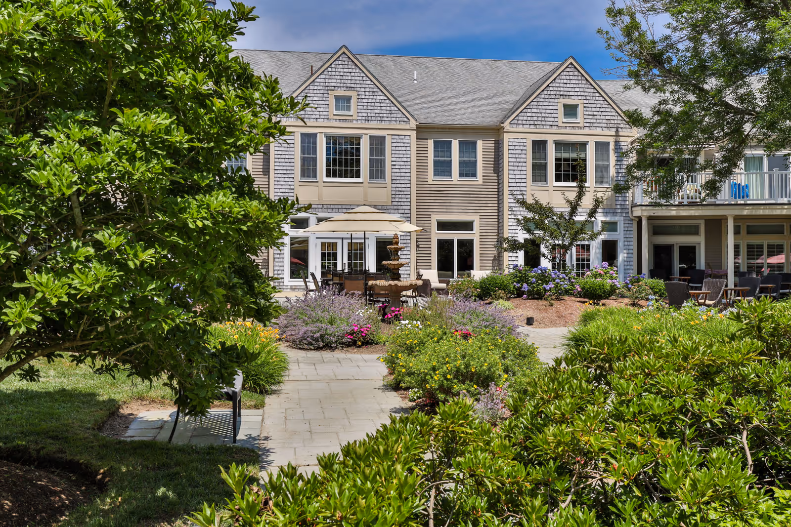 Outdoor garden area at Heatherwood At Kings Way featuring a paved walkway leading to a patio with tables, chairs, and an umbrella. The garden is lush with green shrubs, flowering plants, and trees, with a multi-tiered fountain in the center. The background shows a two-story building with multiple windows and a balcony.