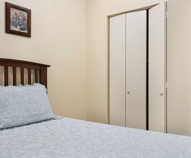 A bedroom with a wooden headboard bed covered with light blue patterned bedding. There is a framed floral painting on the beige wall above the bed and a white closet with double doors partially open.