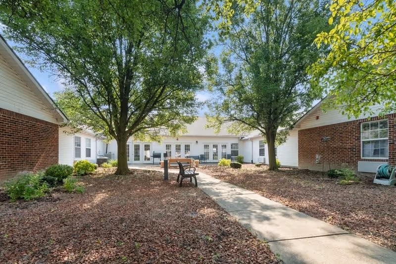 Outdoor courtyard area at Five Star Residences of Northwoods featuring a paved walkway, benches, two large trees, and surrounding buildings with brick and white siding under a clear blue sky.