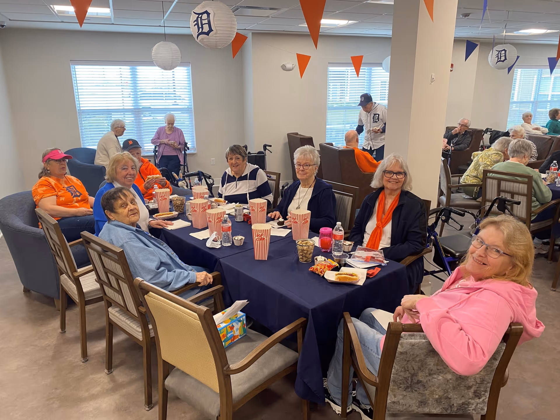 A group of elderly people sitting around a table in a common area, enjoying snacks and drinks. The room is decorated with hanging paper lanterns and triangular pennants in orange and blue. Some people are wearing Detroit Tigers apparel, and there are windows letting in natural light.