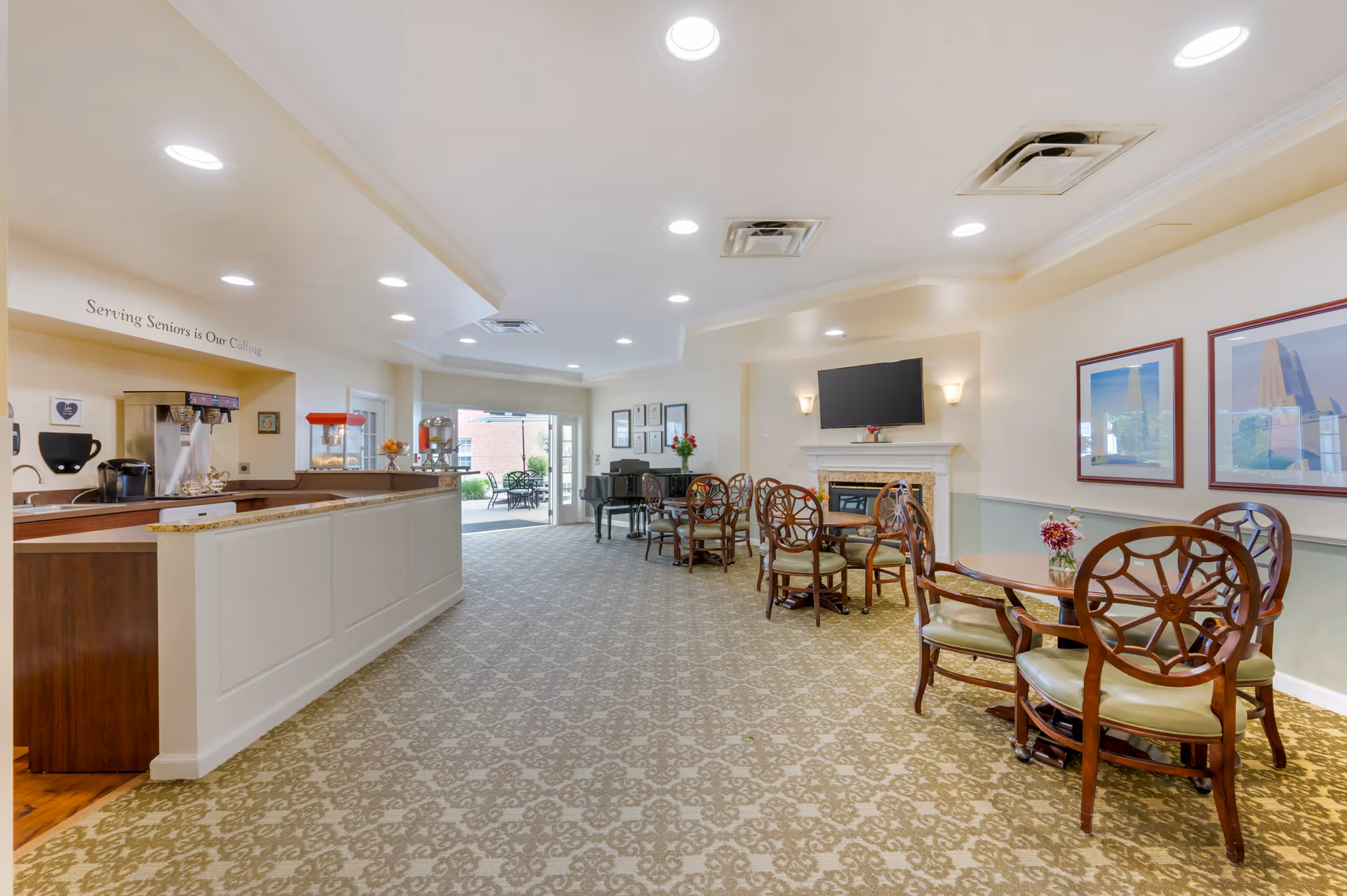 Bright common lounge and dining area with a service counter to the left, round wooden tables and chairs, a TV above a fireplace, and doors leading to an outdoor patio.