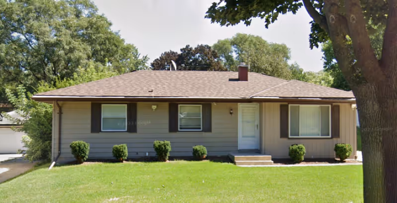 Single-story ranch-style house with a front lawn, three windows, and a centered front door under a brown shingled roof.
