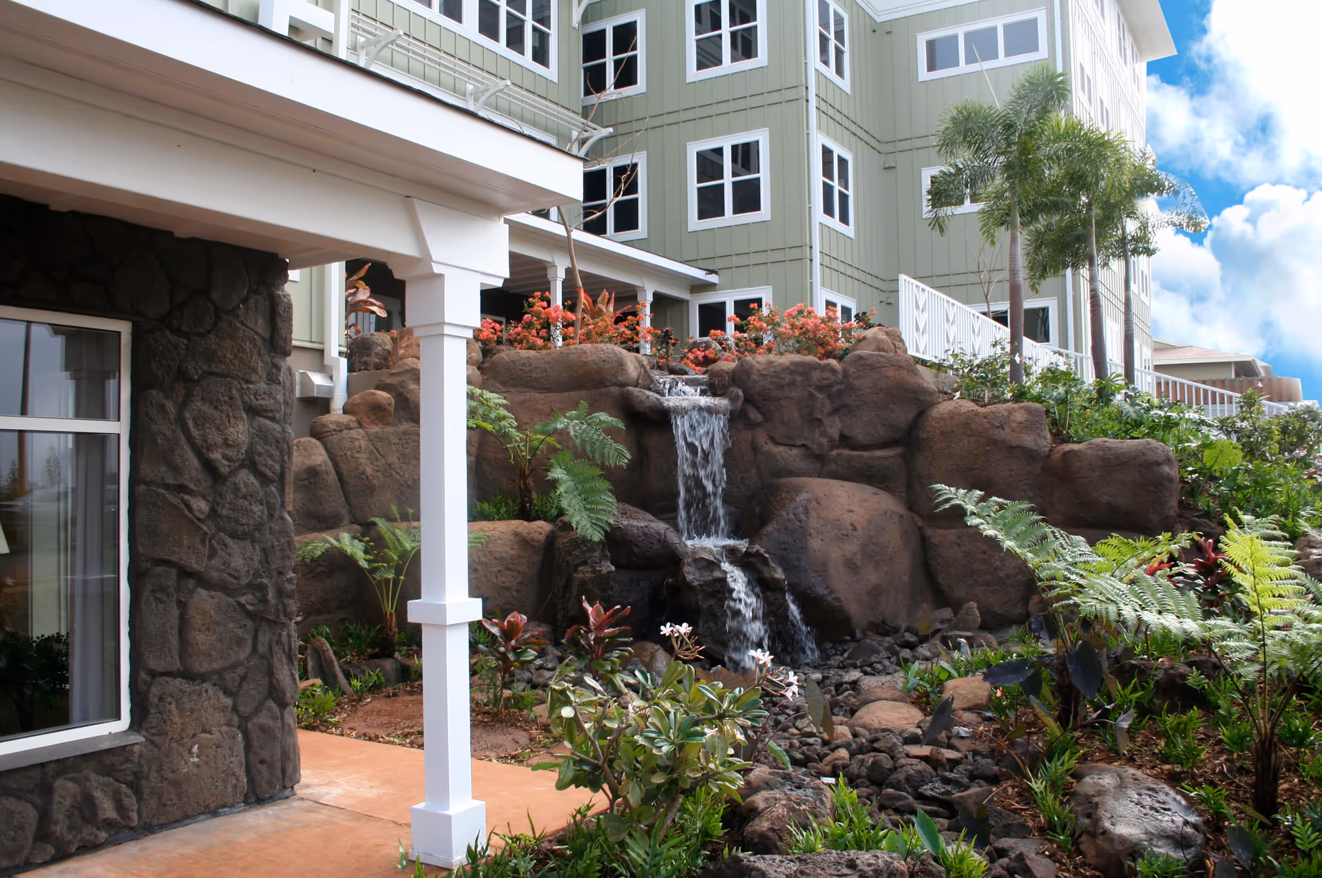 Outdoor garden area at The Plaza at Mililani featuring a small waterfall cascading over large rocks surrounded by lush tropical plants and flowers, with a multi-story green building in the background under a partly cloudy sky.