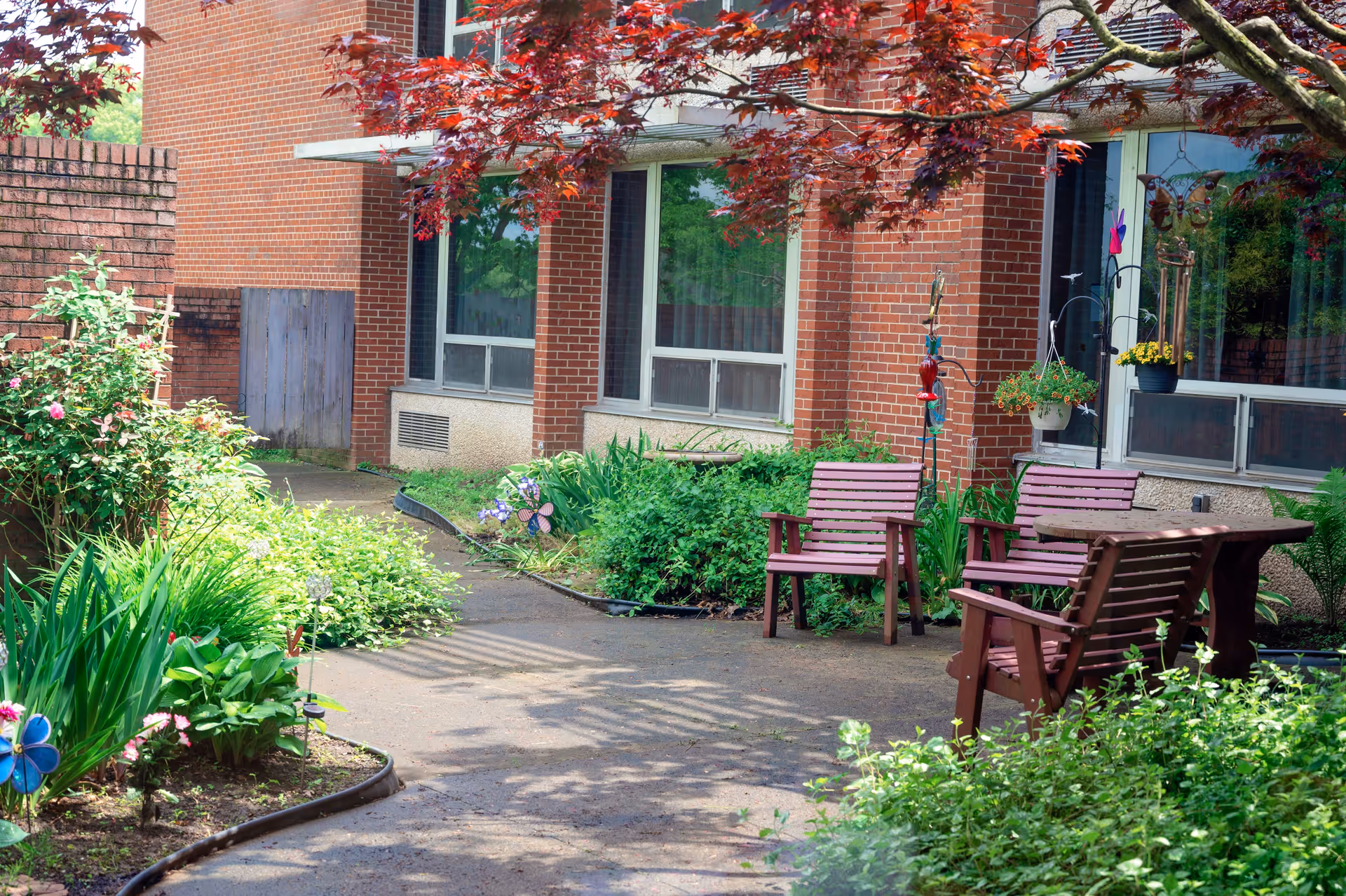 Outdoor garden area with a paved pathway, surrounded by green plants and flowers. There are wooden chairs and a table under a tree with red leaves, next to a brick building with large windows.
