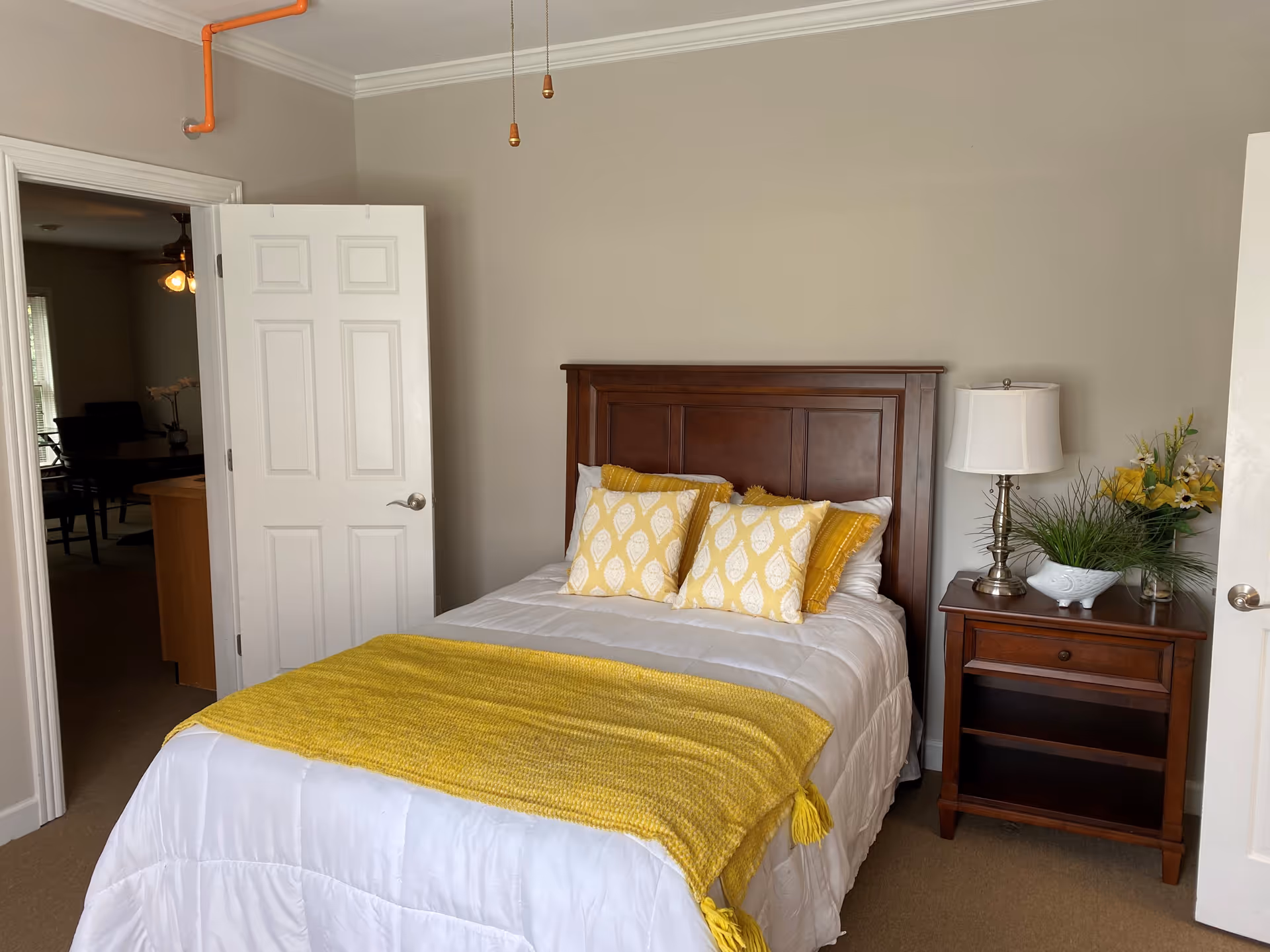 Neatly made bed with yellow pillows and throw, wooden headboard and nightstand with a lamp in a small bedroom.
