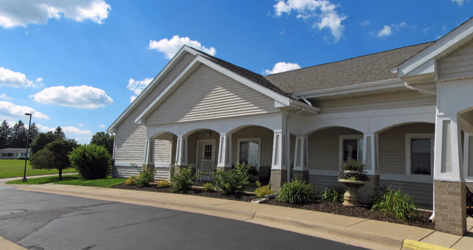 Exterior view of a single-story building with beige siding and white trim under a partly cloudy blue sky. The building has a covered entrance with columns and landscaping including bushes and a decorative planter. A paved driveway and sidewalk are visible in front.