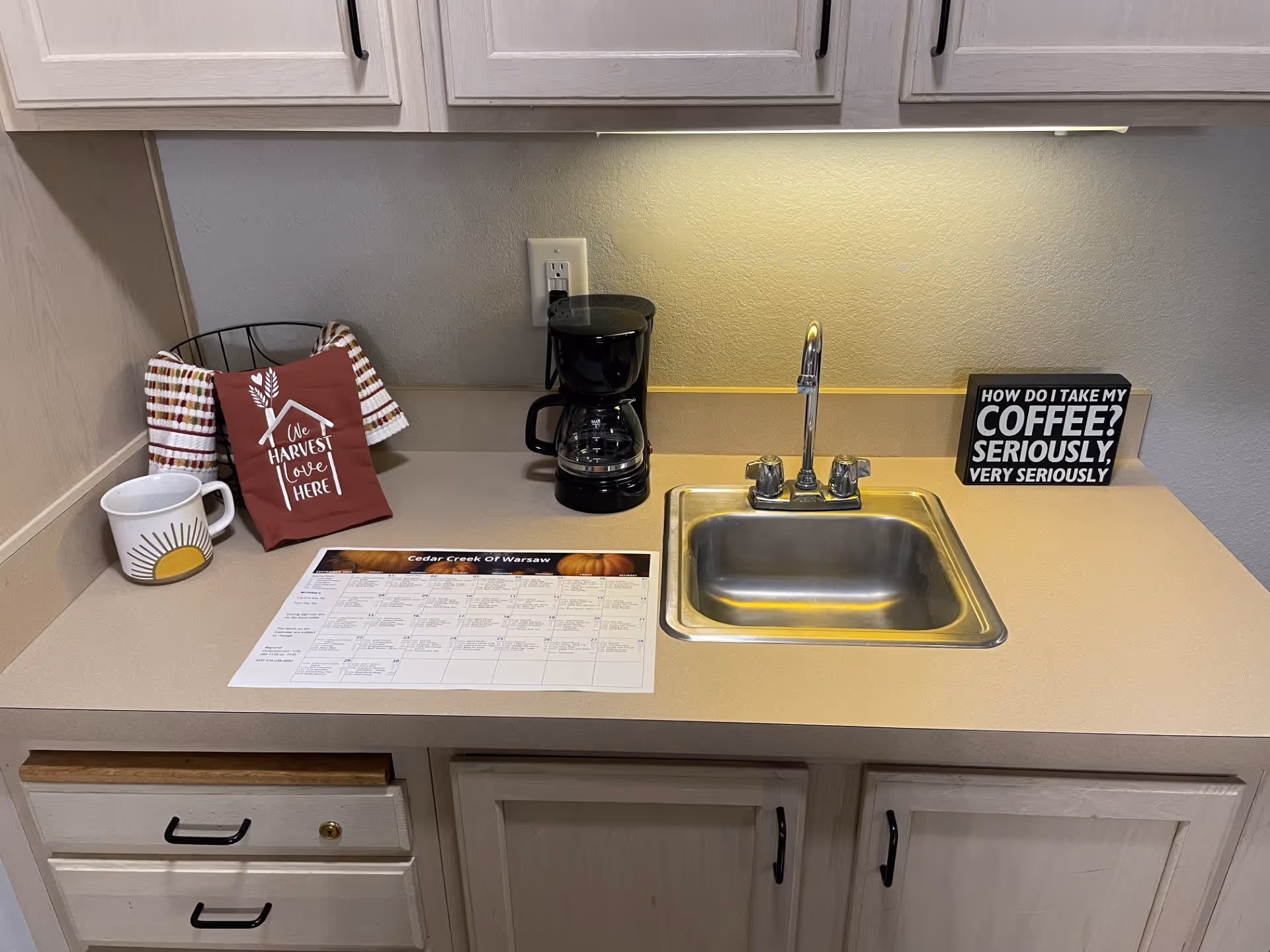 A kitchen countertop with a small stainless steel sink and faucet. On the left side, there is a coffee mug with a sun design, a wire basket holding two kitchen towels and a red towel with the text 'One Harvest Love Here'. In the center, there is a black coffee maker and a printed schedule labeled 'Cedar Creek Of Warsaw'. On the right side, a small sign reads 'How do I take my coffee? Seriously, very seriously'. Light-colored cabinets are above and below the countertop.