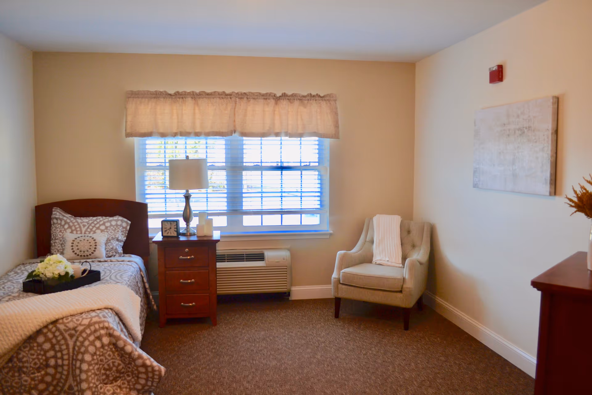 A cozy senior living bedroom with a single bed covered in patterned bedding and a beige throw blanket. Next to the bed is a wooden nightstand with a lamp, clock, and candles. A window with blinds and a beige valance lets in natural light. There is a comfortable upholstered armchair with a white throw draped over it, a piece of abstract wall art, and a wooden dresser with a vase of flowers.