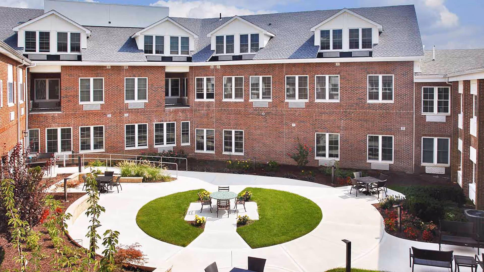 Outdoor courtyard area of a senior living facility with a circular concrete walkway surrounding a grassy center with a table and chairs. The courtyard is bordered by a three-story brick building with multiple windows and dormer windows on the roof. There are additional tables and chairs around the walkway and landscaped plants and shrubs along the edges.