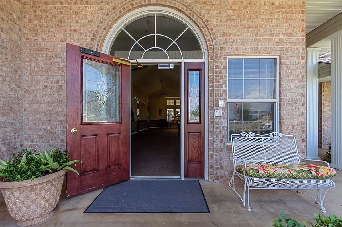 Entrance to a senior living facility with an open wooden door featuring decorative glass panels. The exterior wall is made of light brown brick, and there is a large potted plant on the left side of the door. On the right side, there is a white metal bench with a floral cushion placed under a window with white blinds. The floor is concrete with a dark gray mat in front of the door.