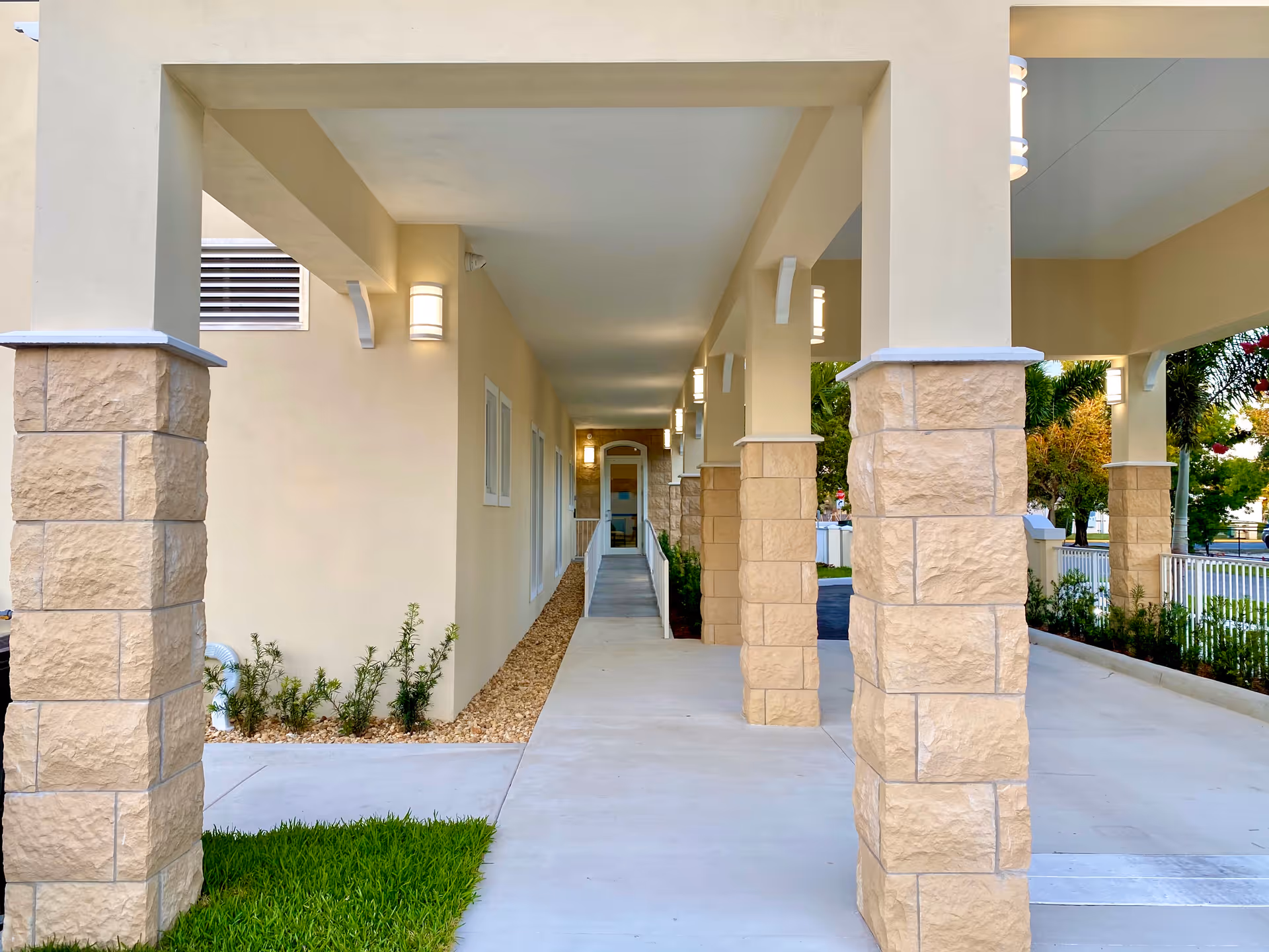Covered walkway with beige stone pillars and cream-colored walls leading to a glass door entrance, surrounded by small plants and greenery.