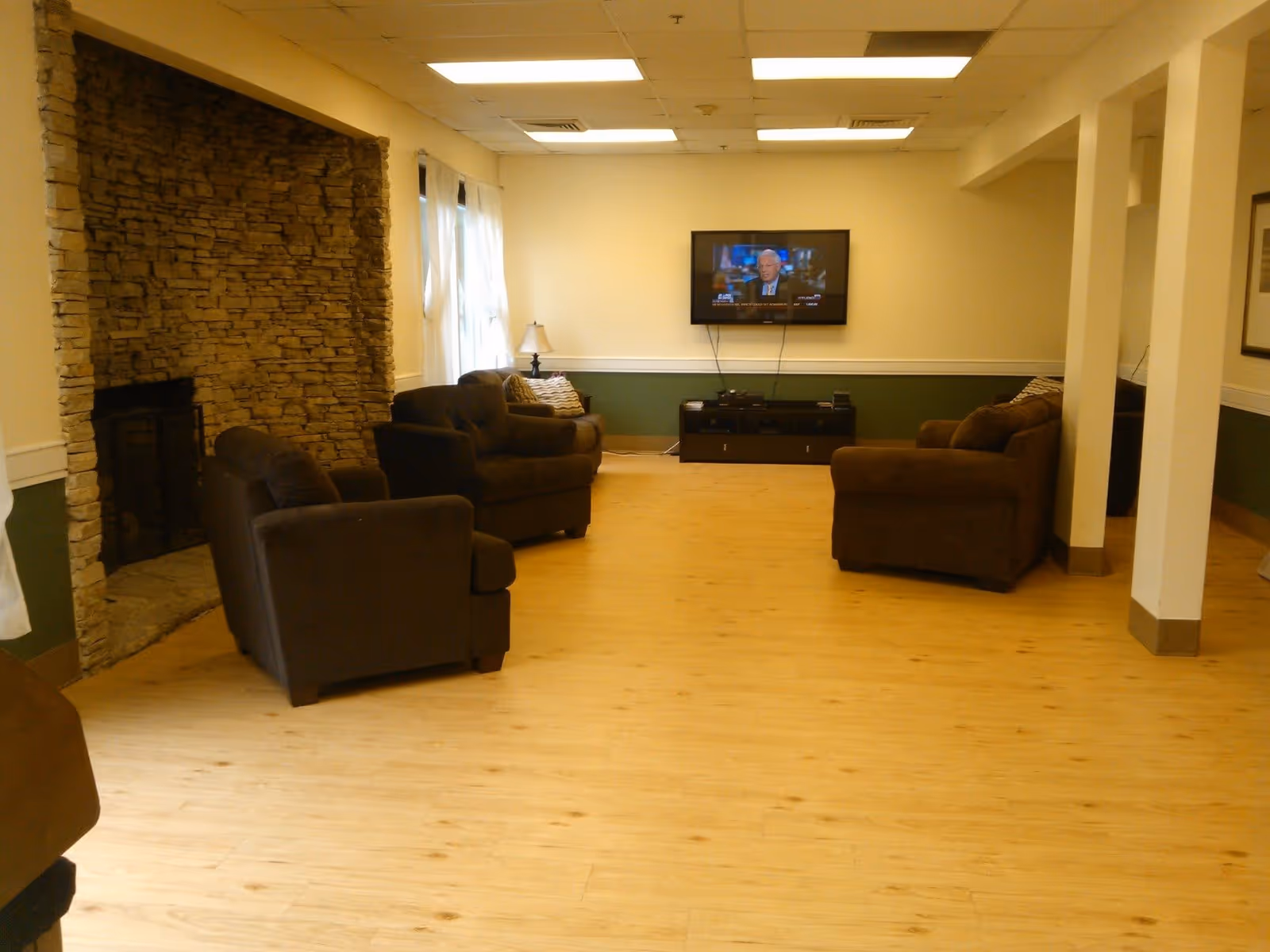 A cozy living room with light wooden flooring, brown upholstered armchairs and sofas arranged around a stone fireplace on the left. A flat-screen TV is mounted on the far wall above a dark wooden media console. The walls are painted cream with a green lower half, and there are windows with white curtains on the left side letting in natural light.