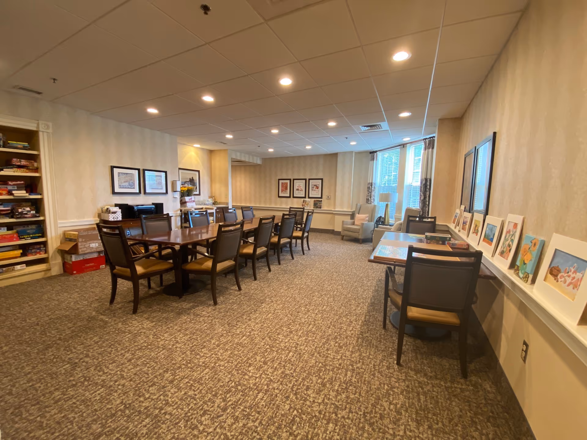 A spacious activity room with a long wooden table surrounded by chairs in the center. On the left side, there is a bookshelf filled with board games and boxes. The right side has smaller tables with chairs and framed artwork displayed on a ledge. The room has beige patterned wallpaper, carpeted floor, recessed ceiling lights, and two large windows with curtains letting in natural light. Two armchairs and a floor lamp are near the windows.